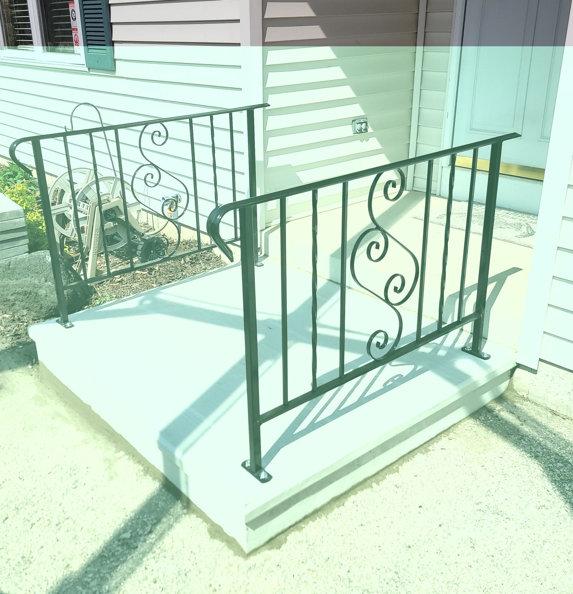 Small front porch with concrete steps and black wrought-iron railing beside a white house.