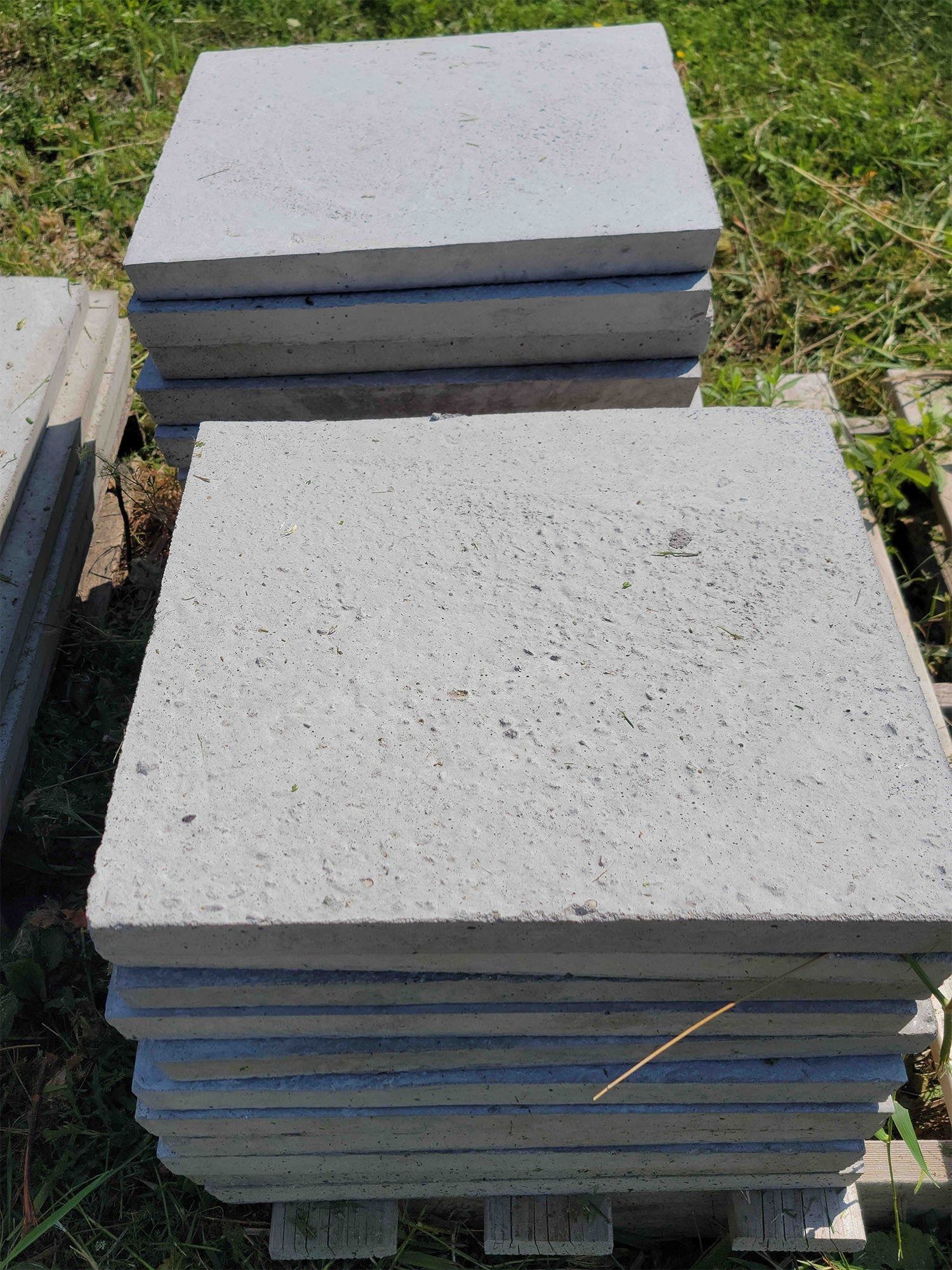 Stacks of gray concrete paving slabs outdoors on a pallet, with grass visible around them.