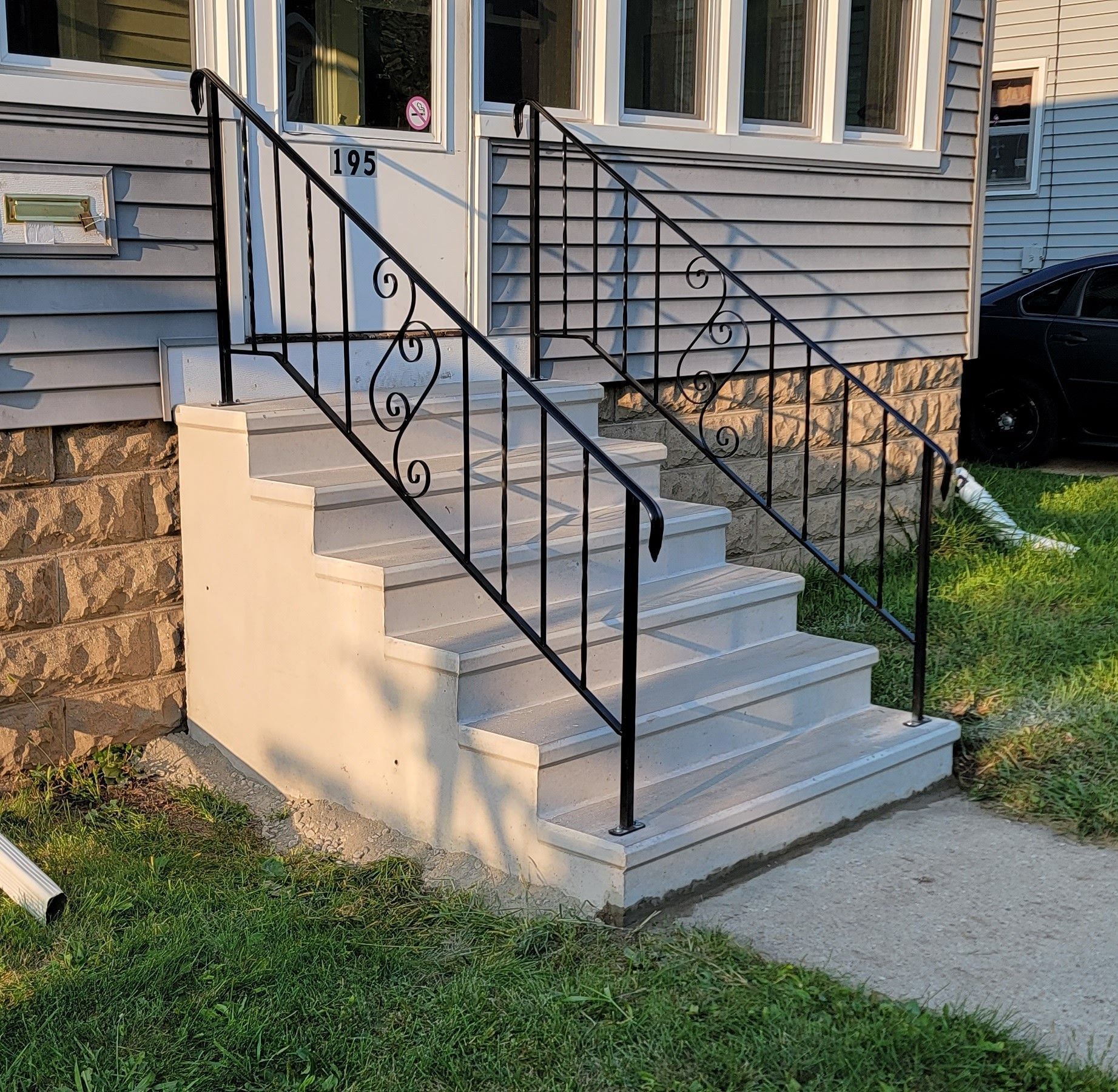 Front steps of a house with white concrete stairs and black metal railing beside a stone-and-siding exterior.