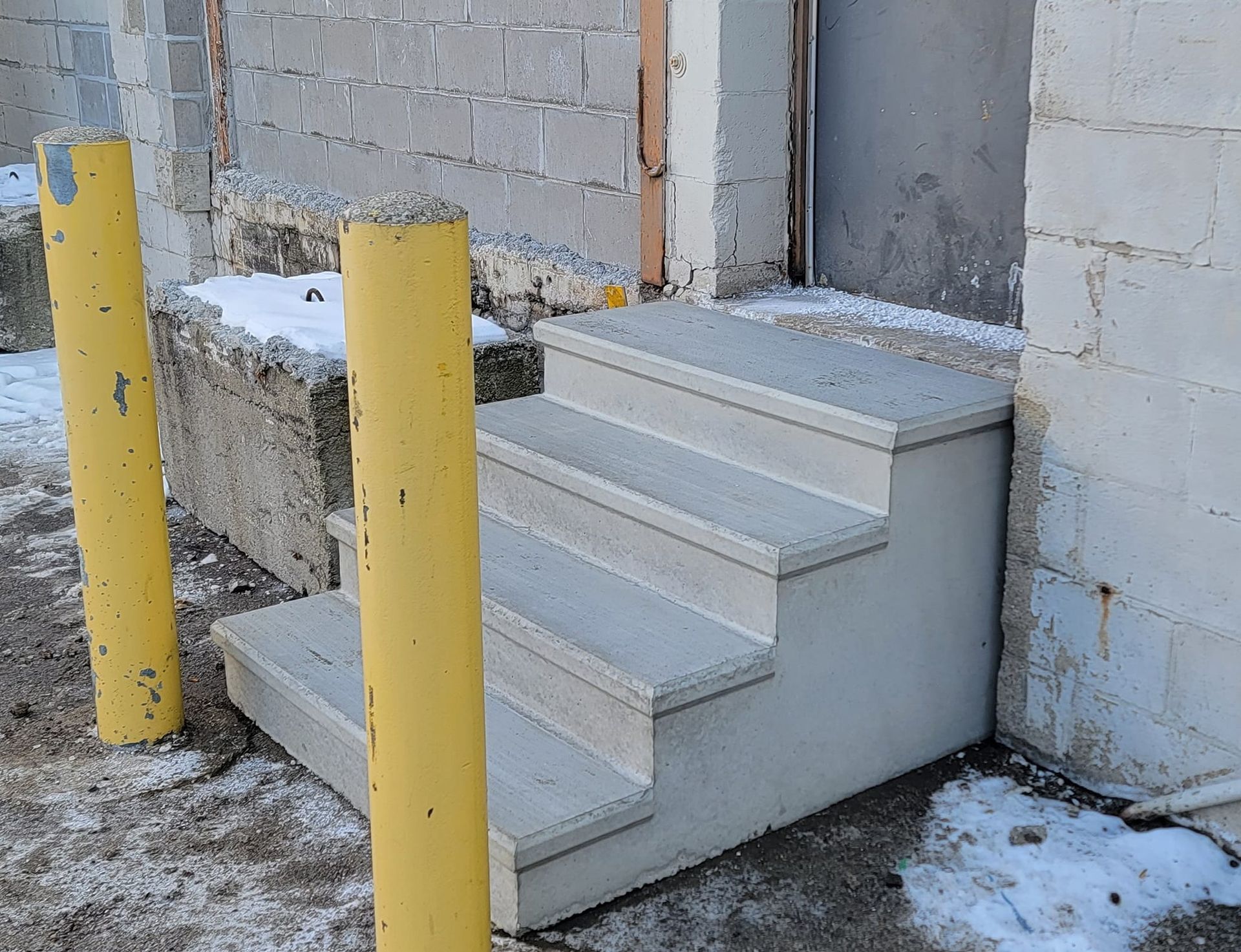 Snowy exterior stairs beside a brick building, with yellow bollards and a gray door.