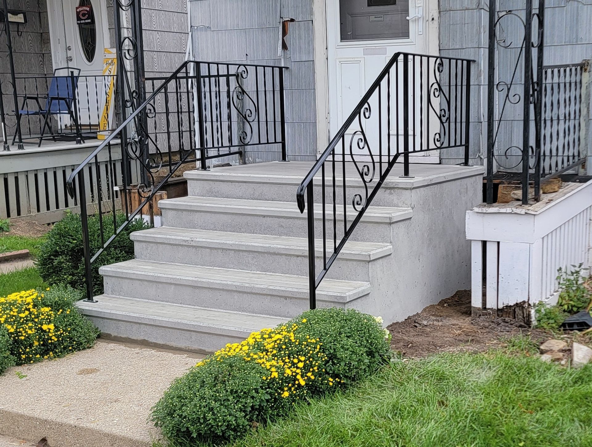 Front steps of a house with black railings, white siding, and landscaped shrubs and flowers