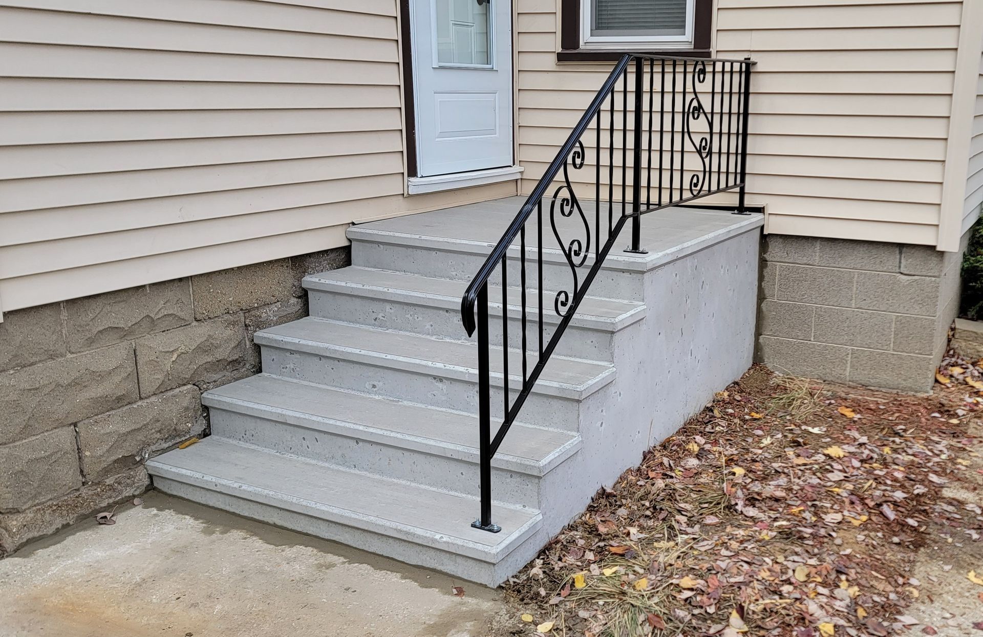 Front steps with gray concrete stairs, black railing, and a white side wall leading to a beige house entrance