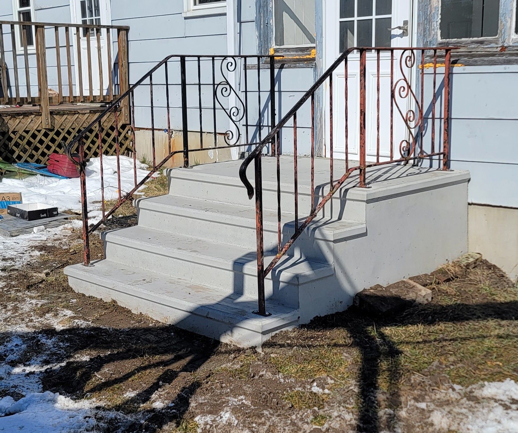 Concrete front steps with black and red railings leading to a house in a snowy yard.