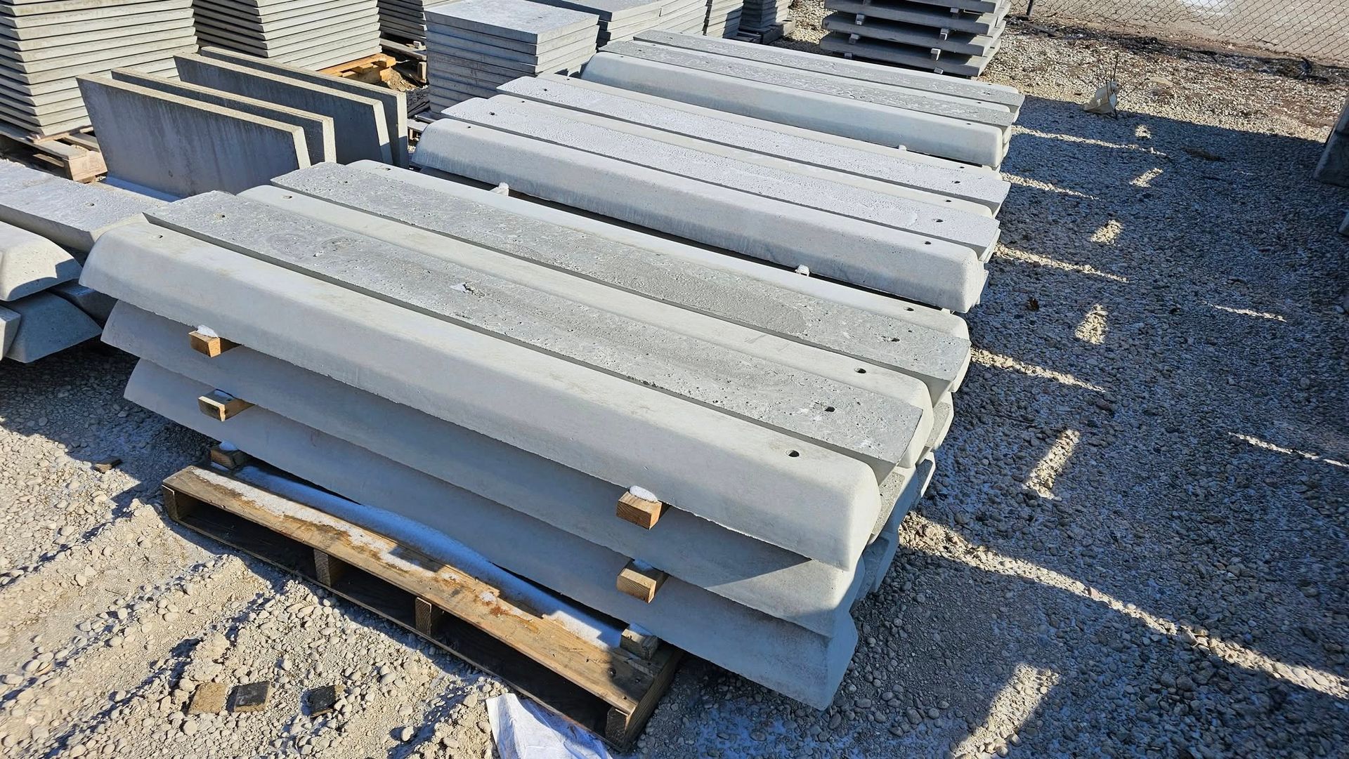 Stacked gray concrete beams on wooden pallets outdoors in a gravel lot.
