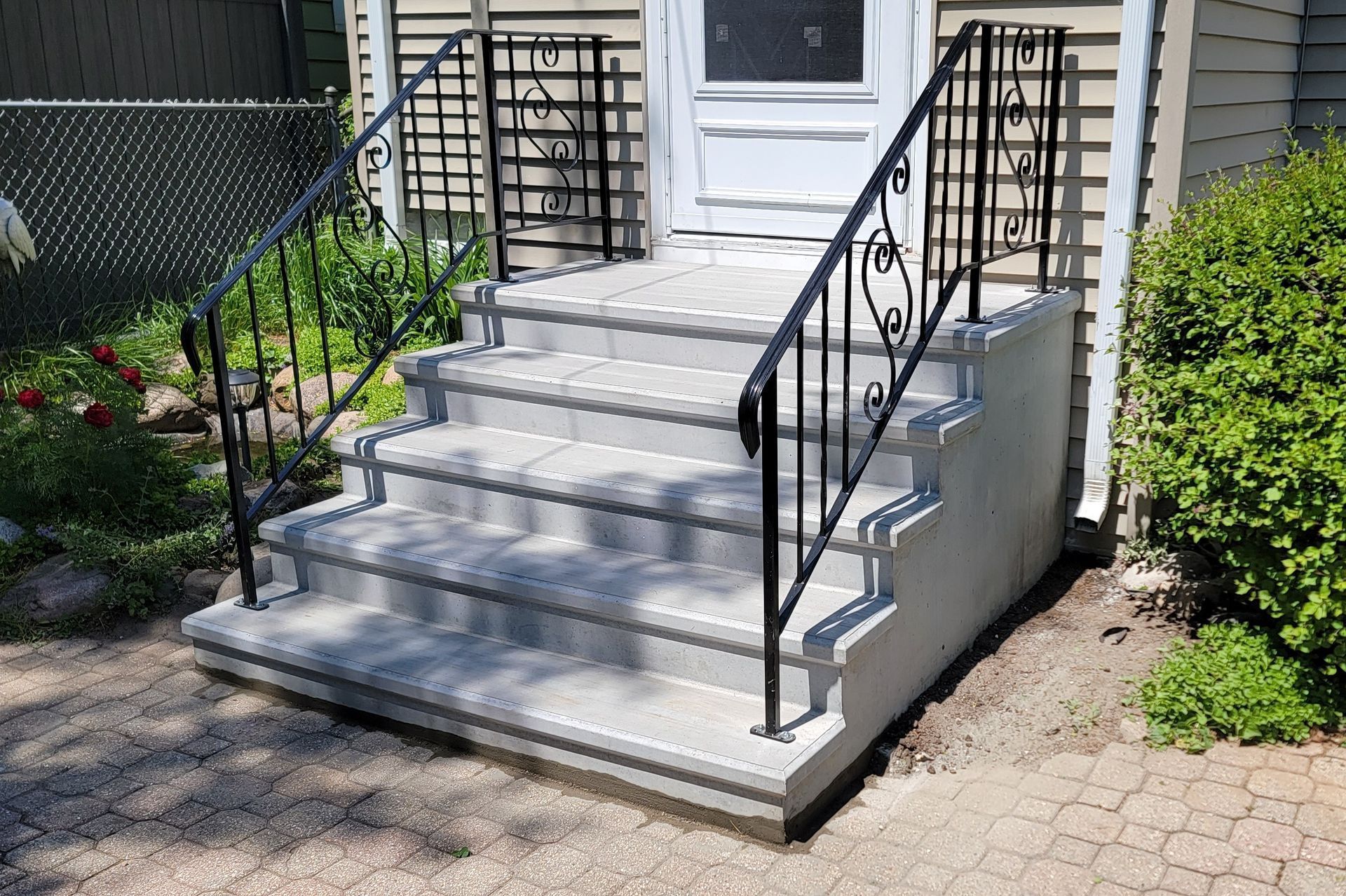 Front steps with black railings leading to a white door, bordered by shrubs and a brick walkway.