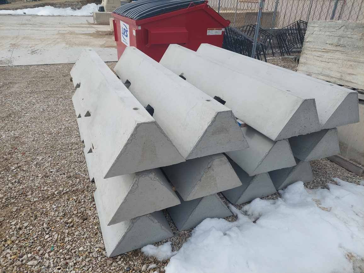 Stacked gray concrete barriers beside a red dumpster on snowy ground outside a fence