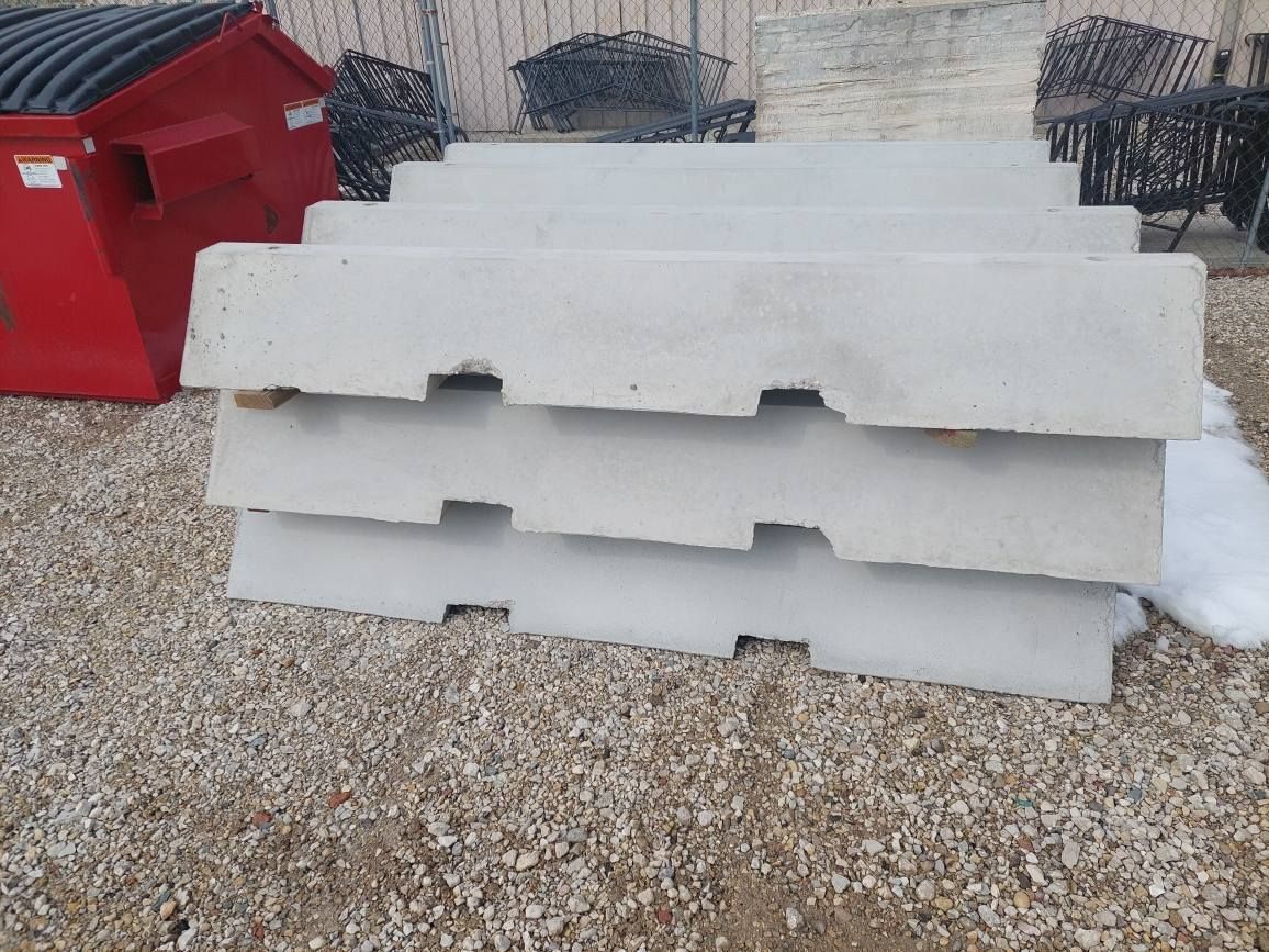 Stacked white concrete barriers on gravel beside a red dumpster and building wall.