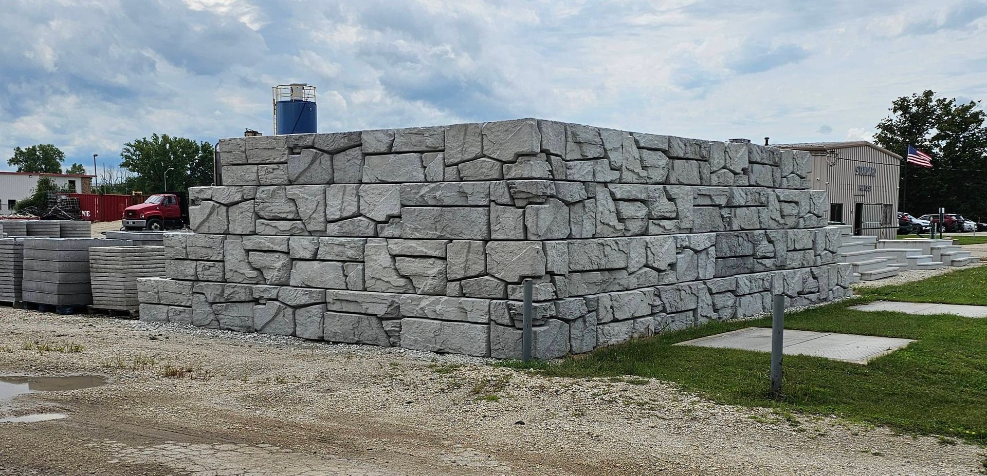 Stacked stone blocks beside a grassy lot under a cloudy sky, with a small tower in the background