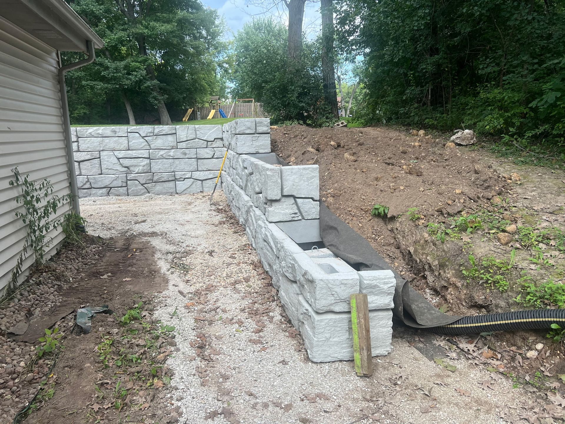 Stone retaining wall and steps beside a house, with gravel path and sloped dirt yard.