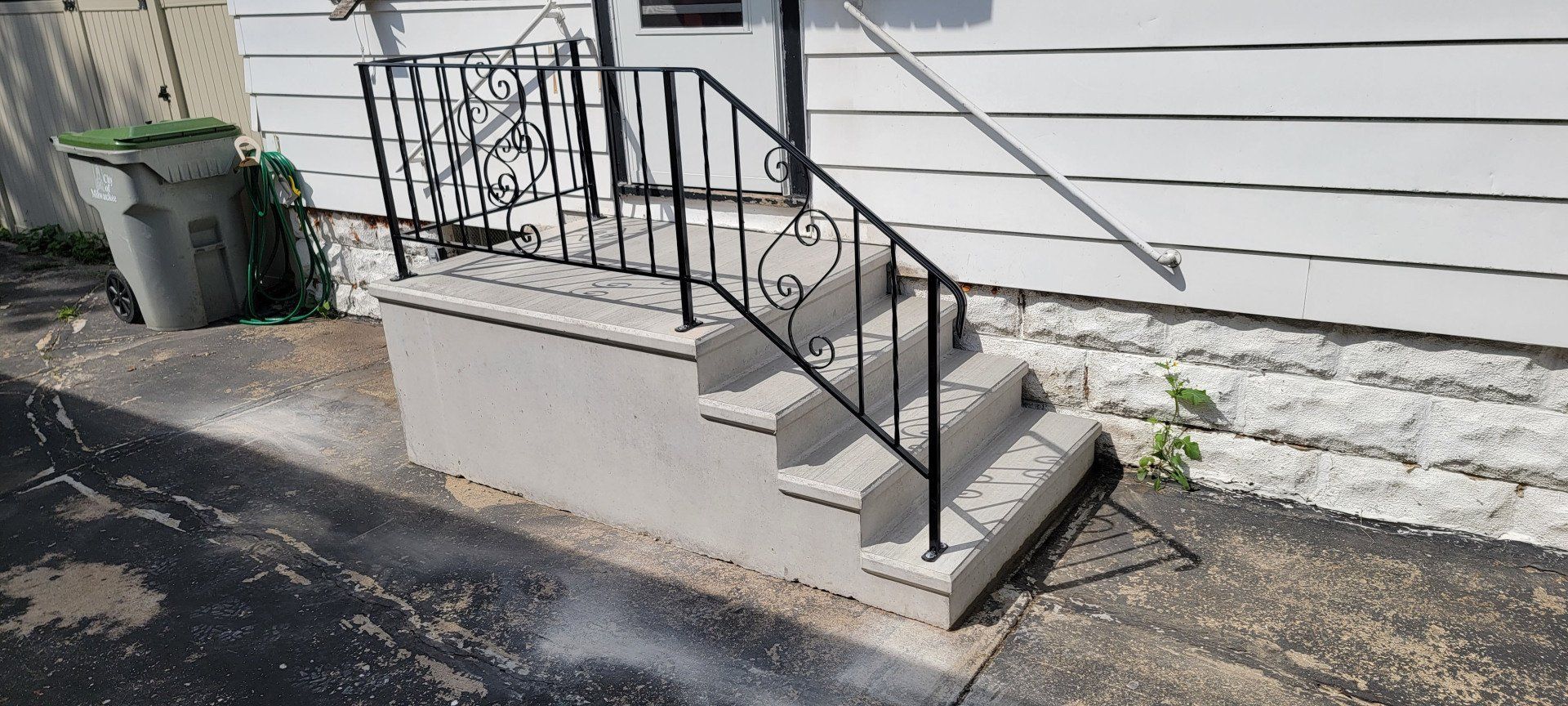 Concrete exterior steps with black handrails beside a white house wall and trash bin