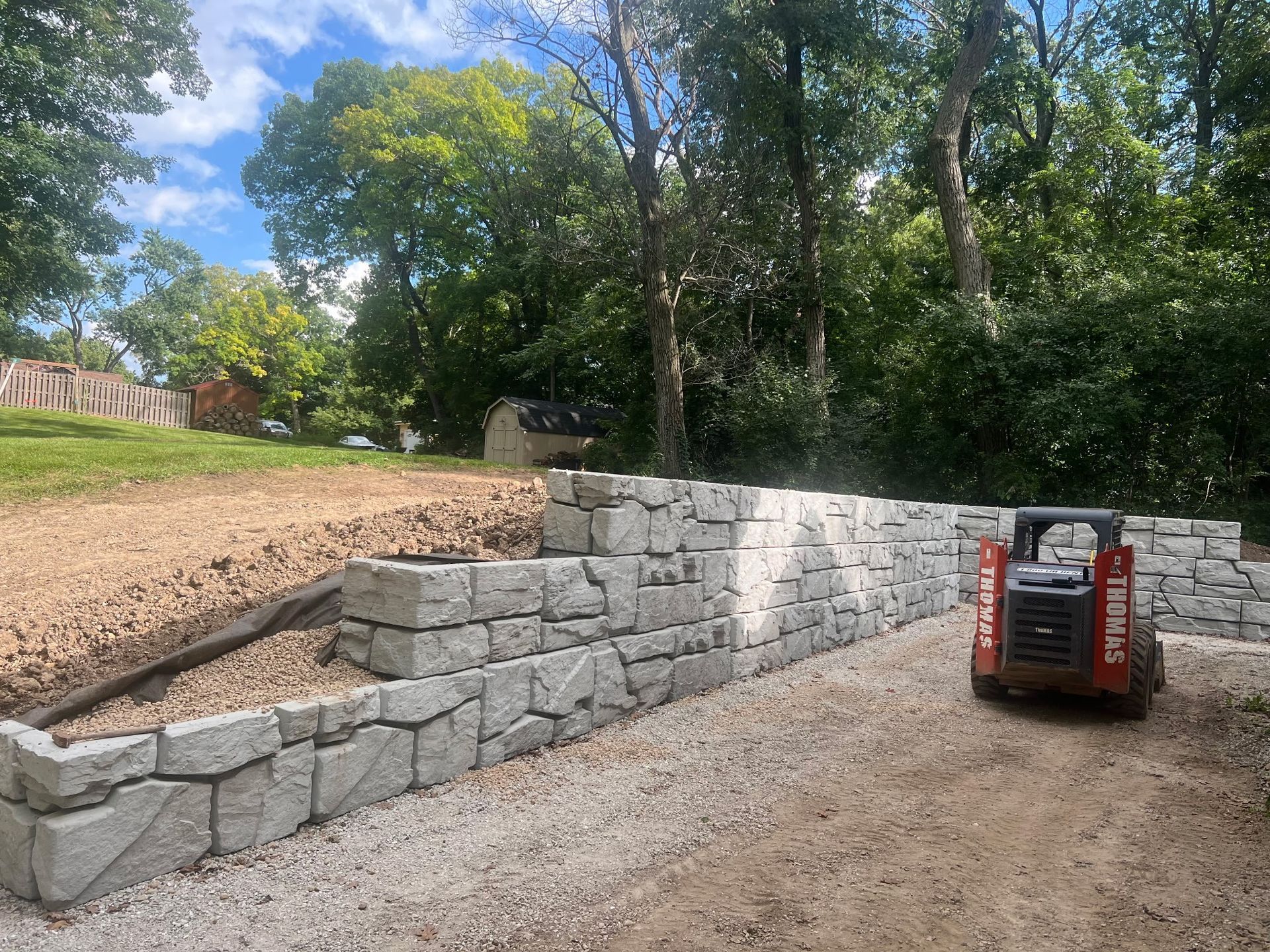 Stone retaining wall under construction beside a gravel driveway, with a red skid steer and trees overhead