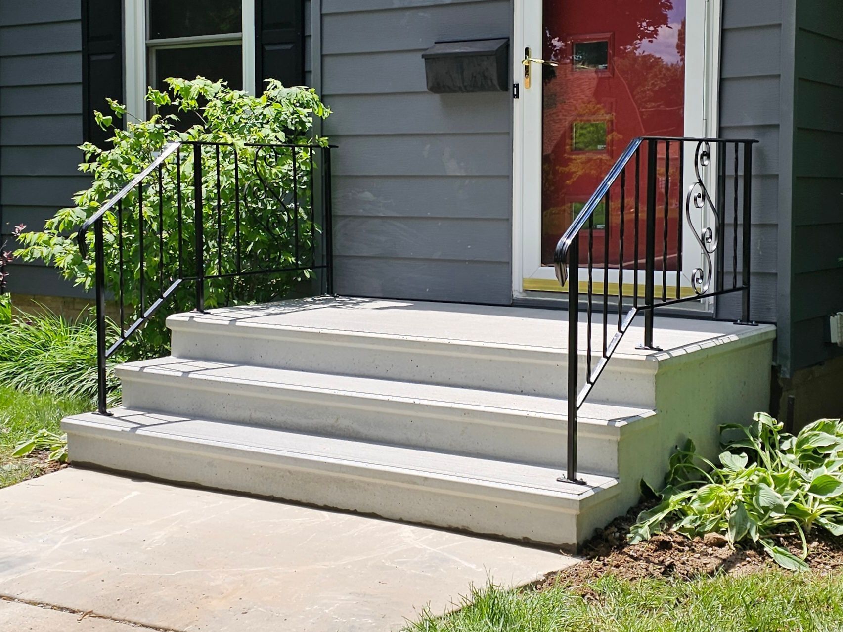 Front steps with black railings leading to a gray house and red door, beside a small garden.