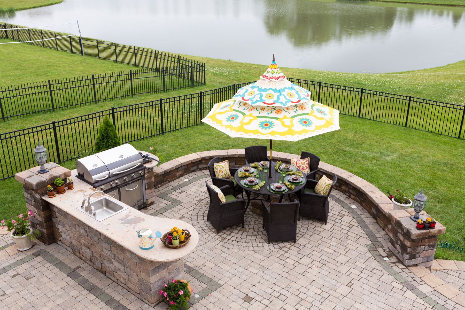 Outdoor kitchen and dining area by a pond, featuring grill, seating, umbrella, and stone patio.