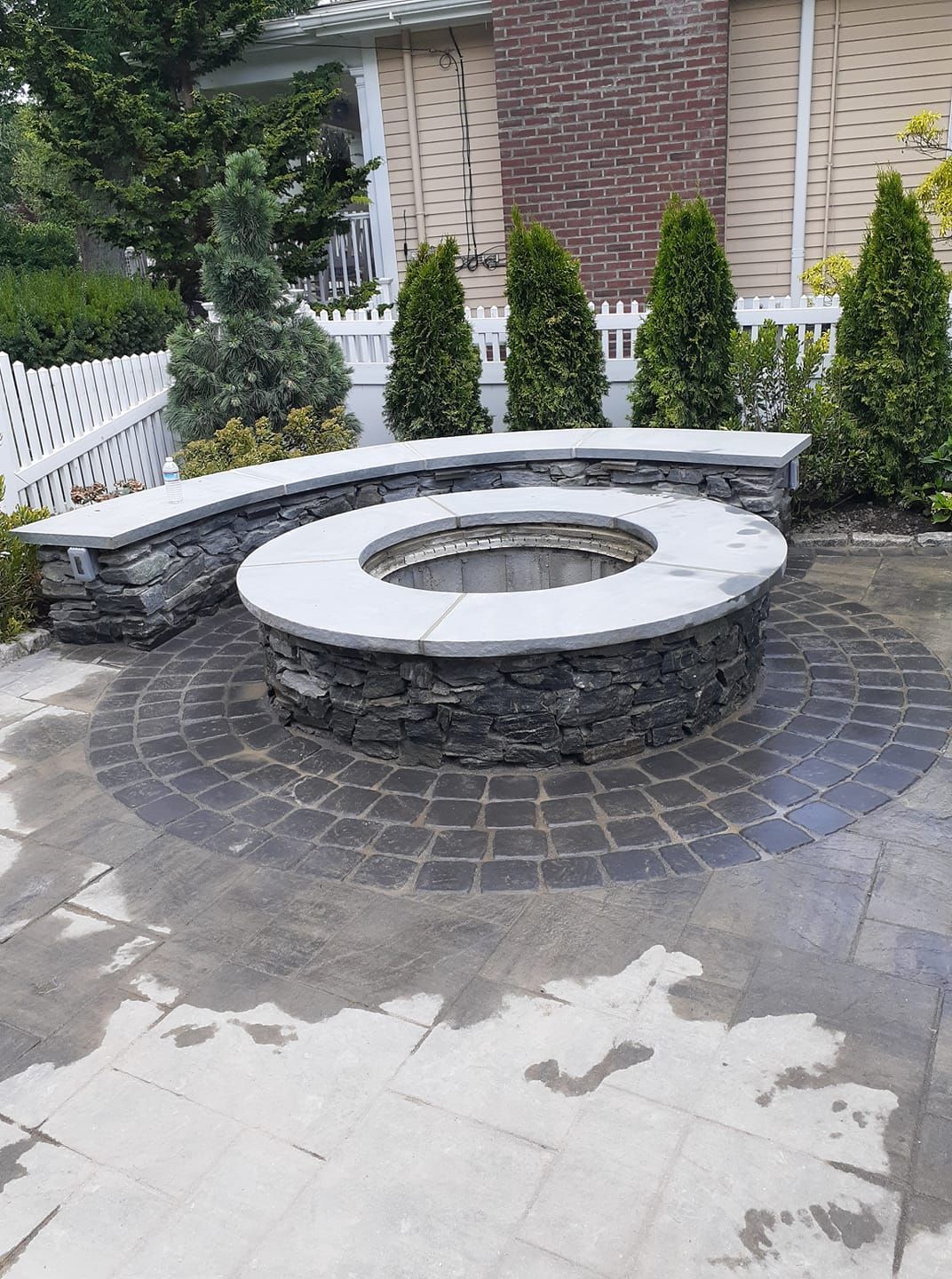 Stone fire pit with curved seating in a backyard. Circular brick pattern and evergreens visible.