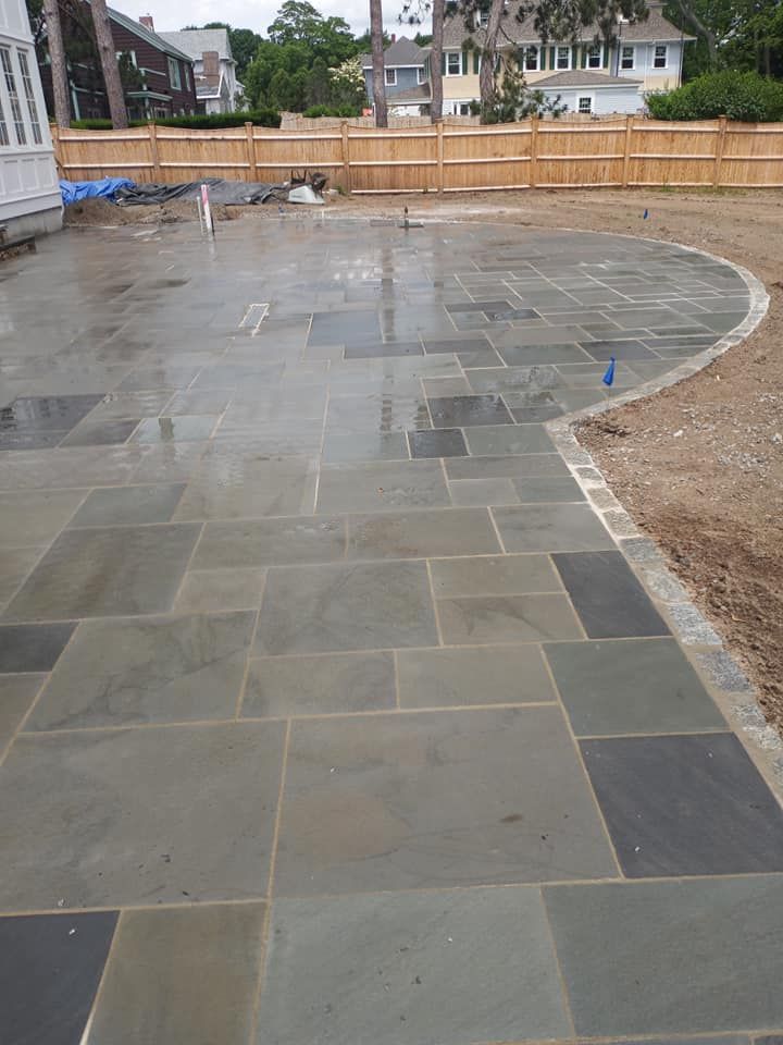 A large patio of blue-gray flagstones with a curved edge, adjacent to a dirt area, with a wooden fence in the background.