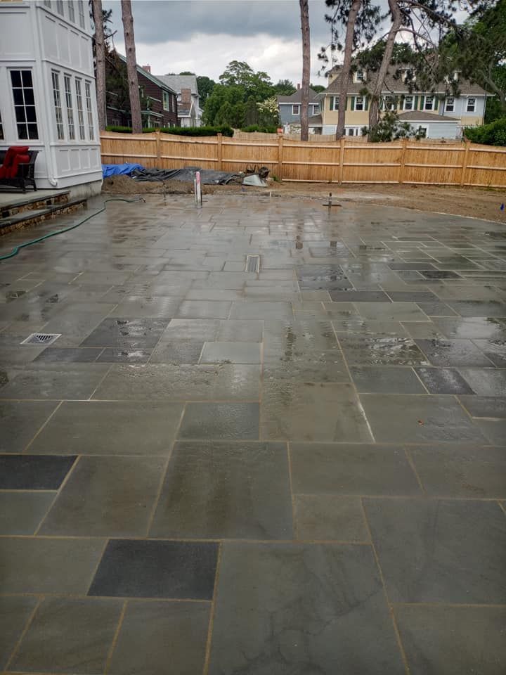 Wet, gray stone patio with various sized rectangular tiles. A wooden fence and houses are in the background.