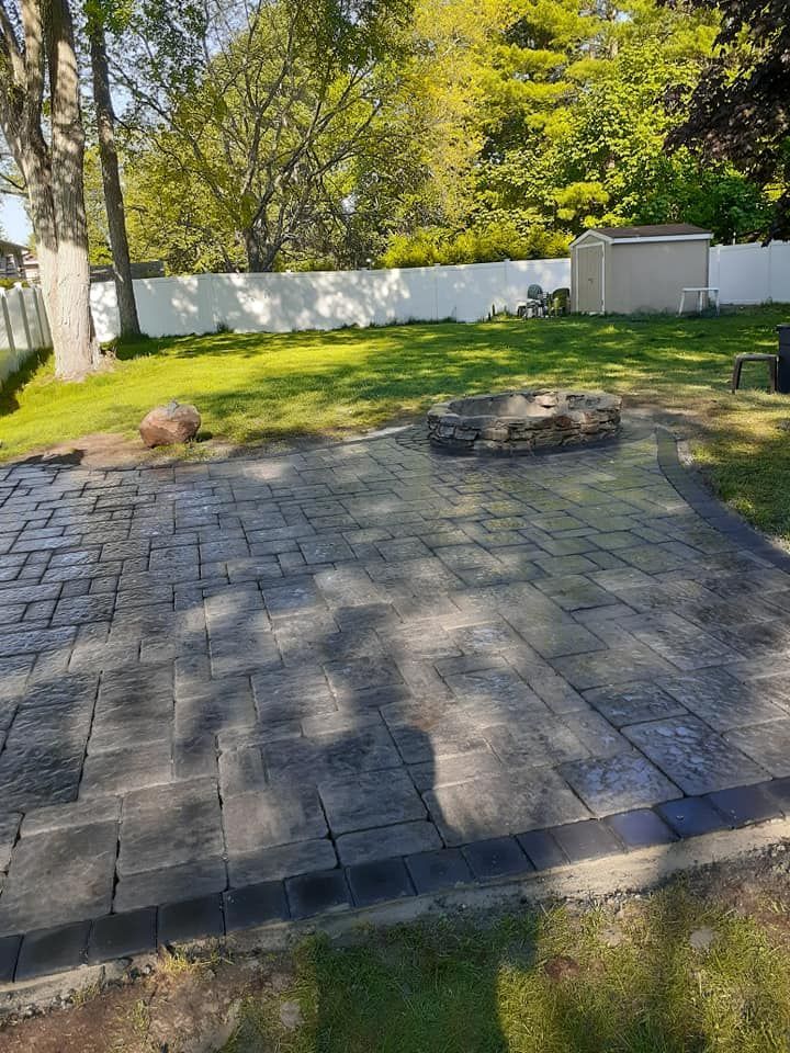 Brick patio with a fire pit in a grassy backyard, surrounded by a white fence and trees.