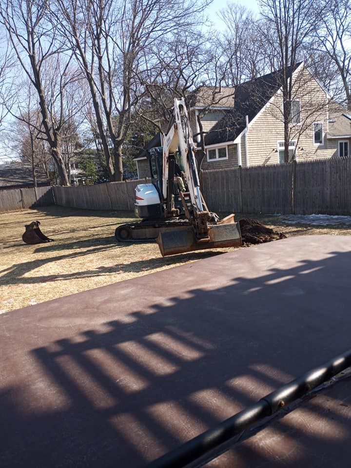 Mini excavator digging in a grassy backyard, brown pavement in foreground, house and fence in background.