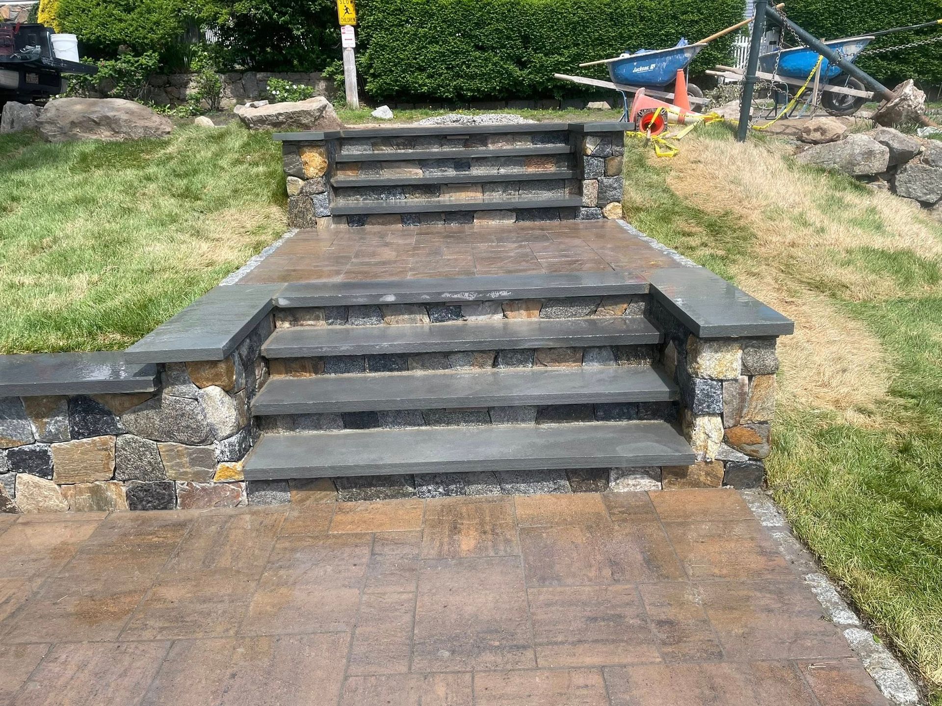 Stone steps leading up to a tiered patio. Brown and gray stonework, surrounded by grass.
