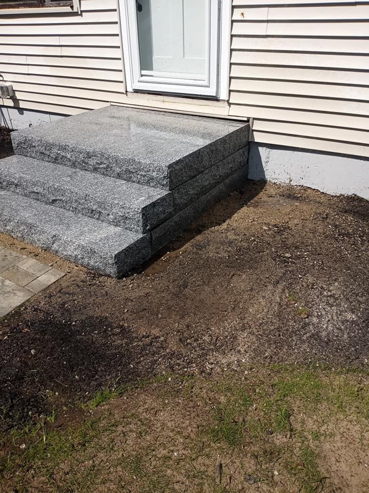 Granite steps leading up to a white door; steps are surrounded by dirt and grass, house is light brown.
