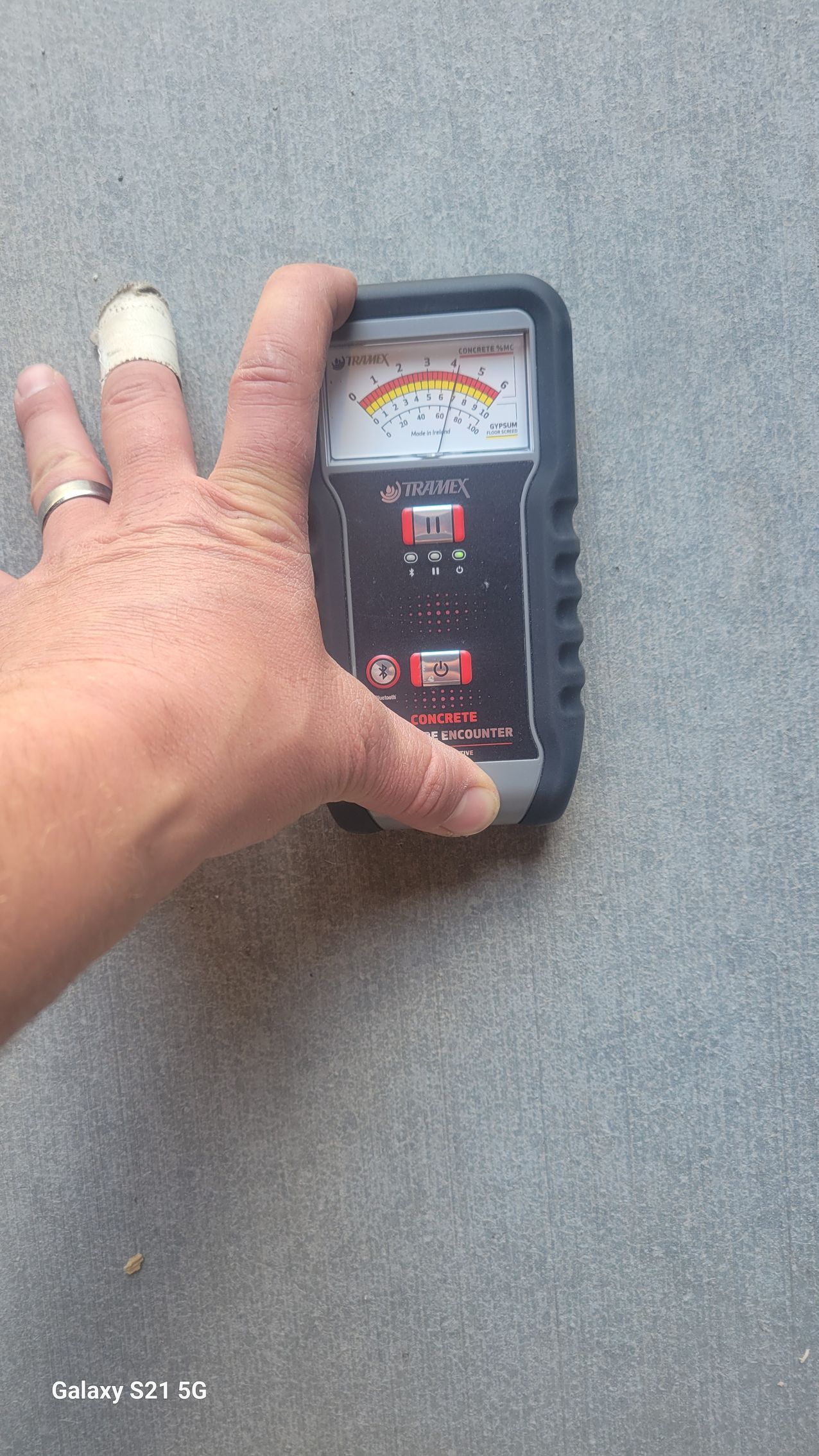 Hand holding a gray handheld meter with an analog display against a gray surface.