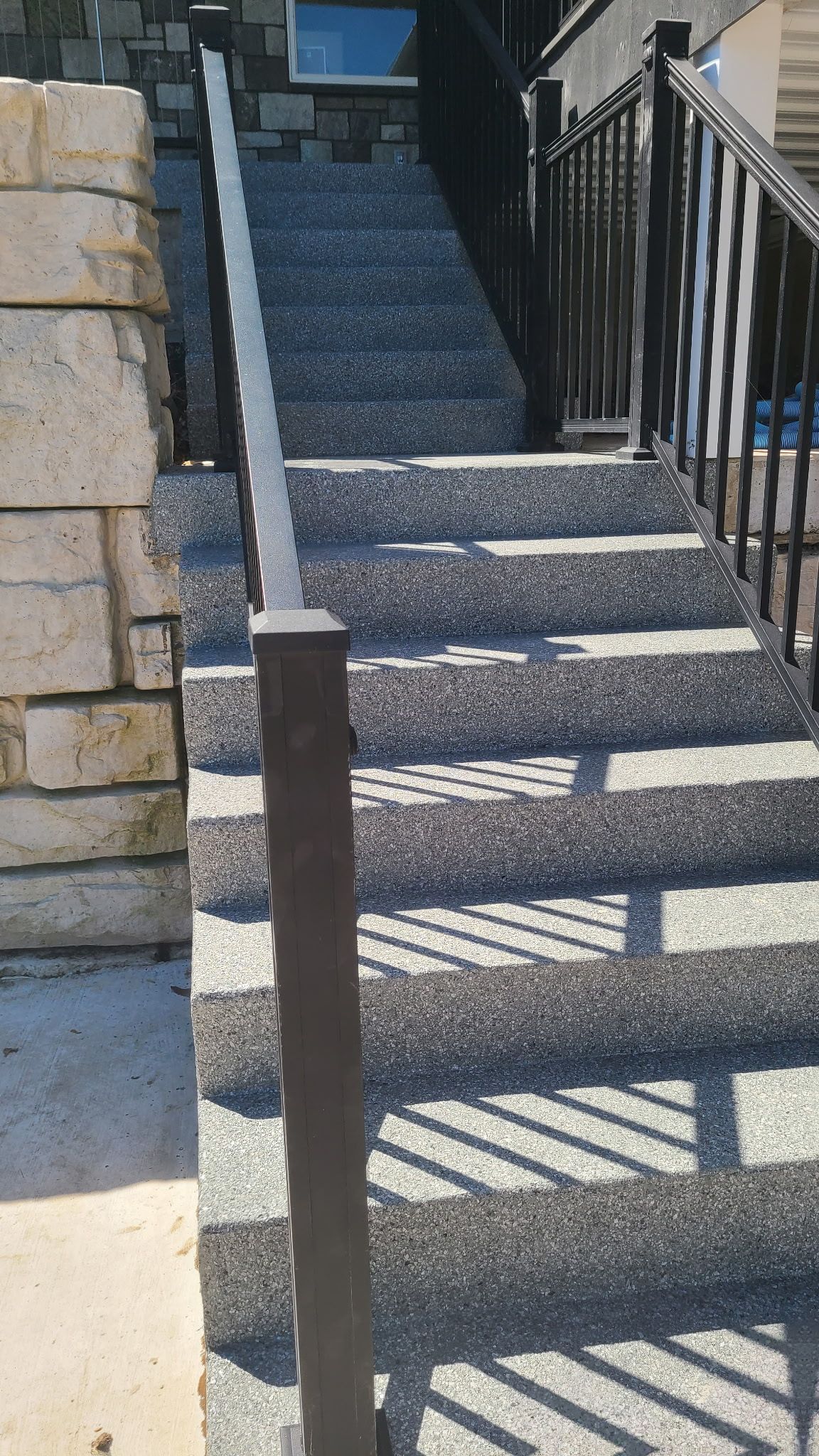 Outdoor concrete stairs with metal railings beside a stone wall, casting striped shadows.