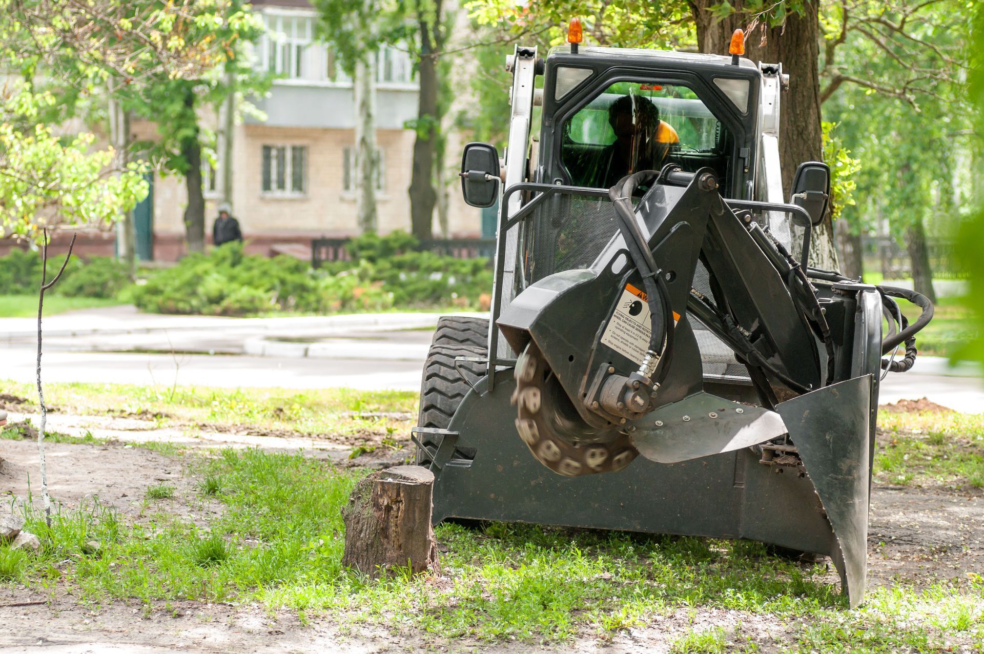 Skid steer grinding a tree stump in a grassy area; building in background.