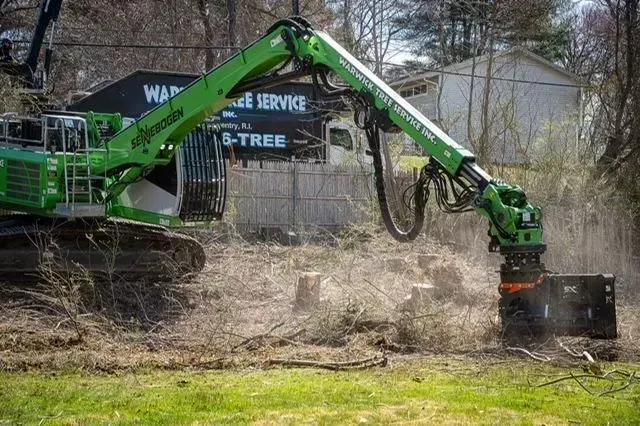 Green tree stump grinder removing stumps in a grassy area, with a white vehicle and a house in the background.