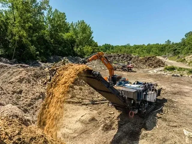 Excavator dumping wood chips into a large screening machine, outdoors in a clearing.