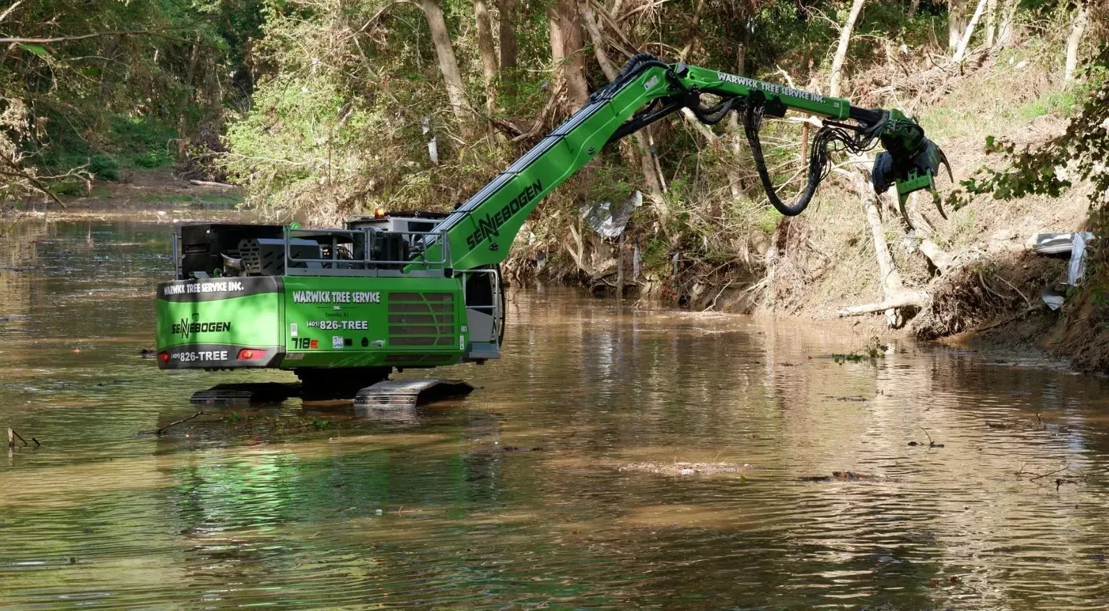 Green excavator dredging murky water in a creek, trees in the background.
