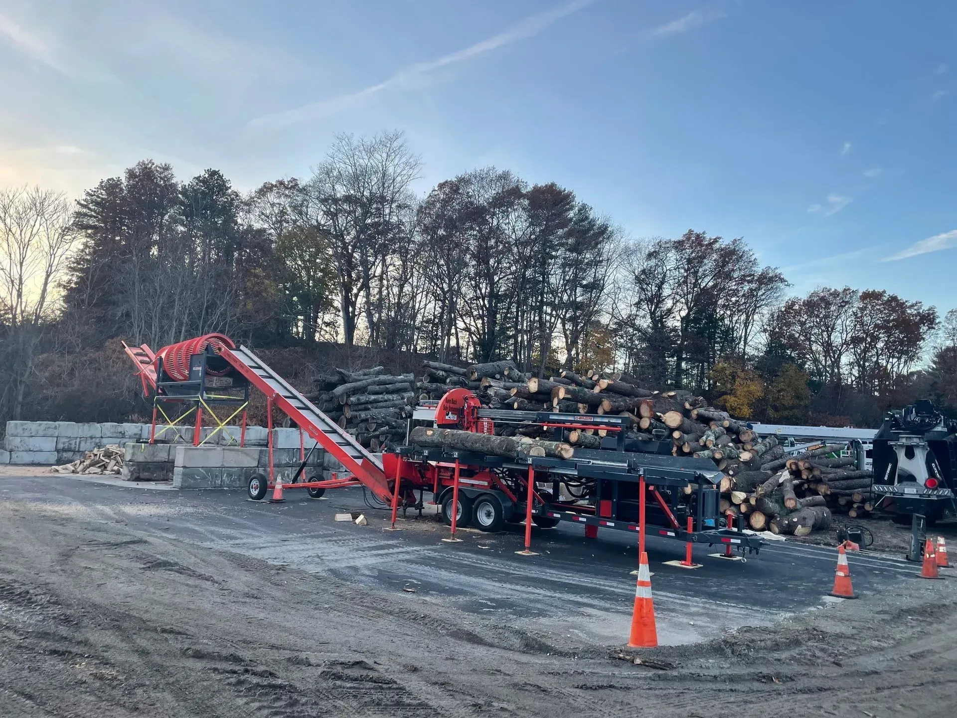 Red wood chipper and conveyor loading logs onto a pile outdoors on a gravel lot; trees in the background.