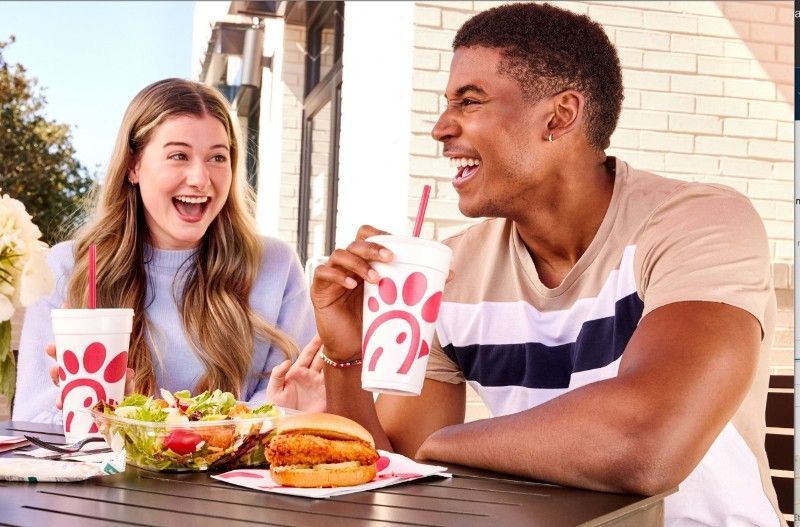 Young couple laughing, eating Chick-fil-A outdoors. Man holds drink, woman eats salad.
