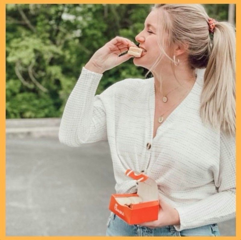 Blonde woman in white sweater eating from a Chick-fil-A box outdoors; smiling.