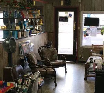 Interior of a small hair salon with wooden paneling. Two wicker chairs face the door; shelves hold products and a hooded dryer stands nearby.