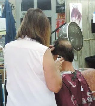 A hairdresser cuts the hair of a client in a salon. The client wears a salon cape and sits under a hair dryer.