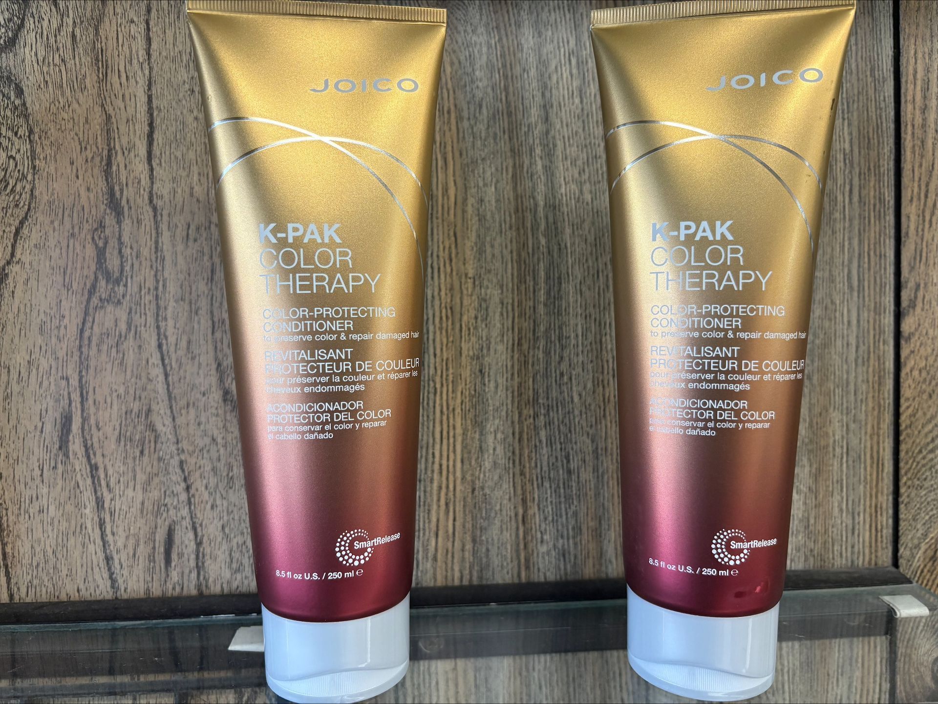 Two tubes of Joico K-Pak Color Therapy hair product, gold and ombre design, on a wood surface.