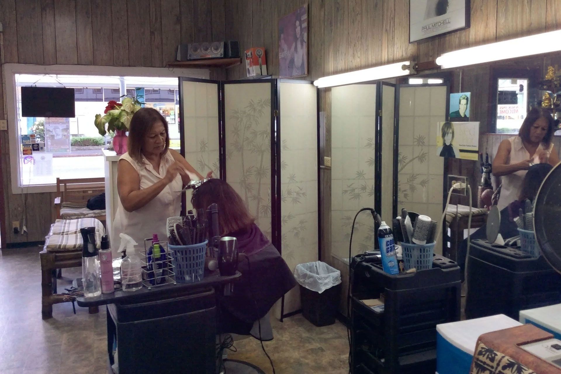 A woman getting her hair styled in a salon. A stylist works on her hair near a room divider. The salon has wood paneling and a large window.