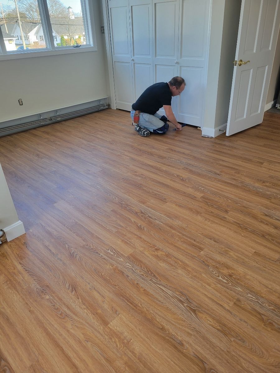 Person installing wood-look flooring near a white cabinet and door in a room.