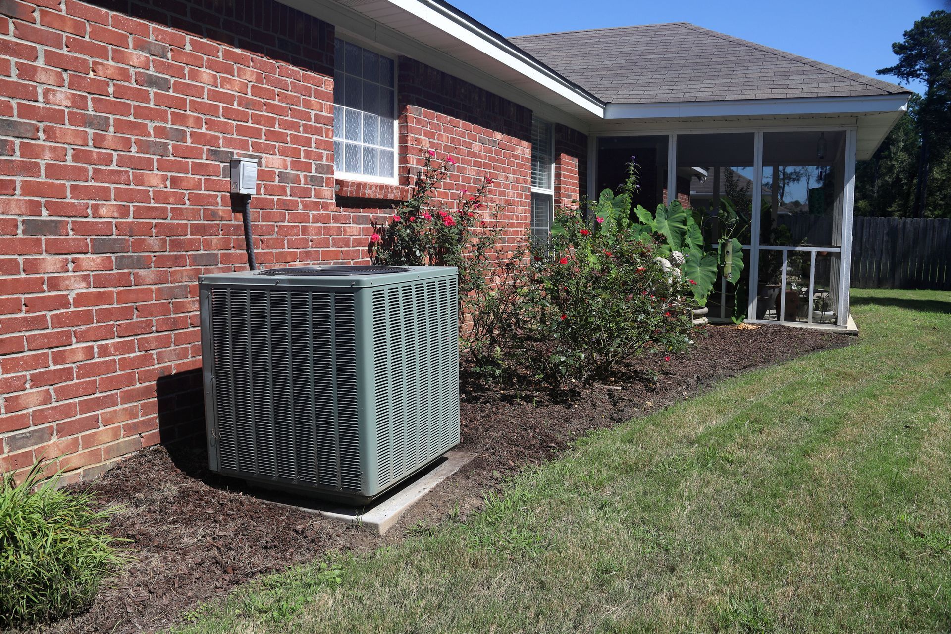 A grey air conditioning unit sits on a concrete pad next to a brick house wall near a screened porch and garden beds.