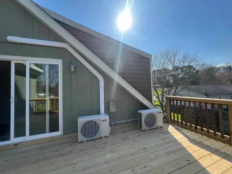 Two HVAC units placed on a wooden deck next to a house exterior with green siding, a glass sliding door, and a railing.