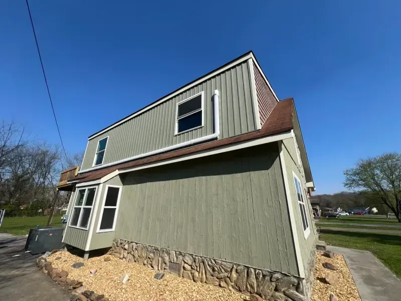 A green, two-story house with stone foundation, bay window, and brown roof under a clear blue sky.