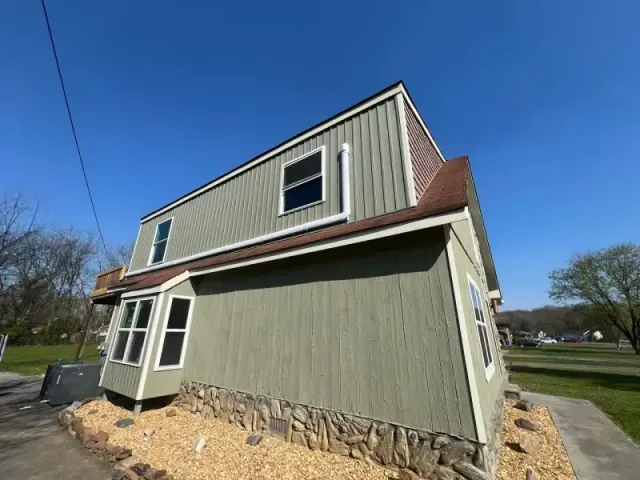 A two-story house with green vertical siding, a stone base, a bay window, and a brown roof against a clear blue sky.