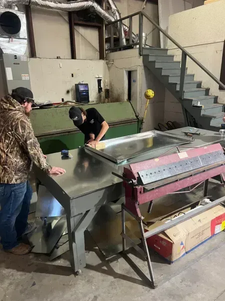 Two workers use a metal brake and table in a workshop to bend a large sheet of metal.