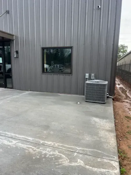An outdoor view of a grey metal industrial building with a window, a concrete patio, and an HVAC condenser unit.