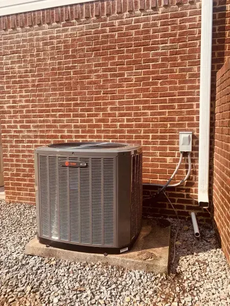 A gray Trane HVAC outdoor unit sits on a concrete pad against a red brick wall, next to an electrical box and drain pipe.