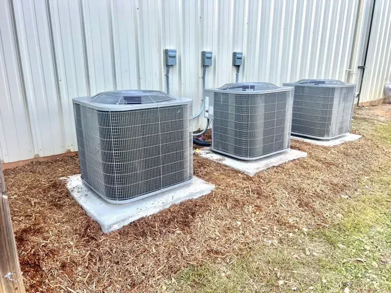 Three gray air conditioning units sit on concrete pads outside a metal-sided building surrounded by wood mulch.