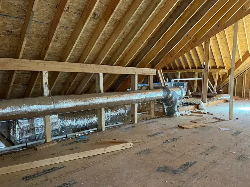 A wooden attic interior showing exposed rafters, a plywood floor, and silver ductwork running horizontally.