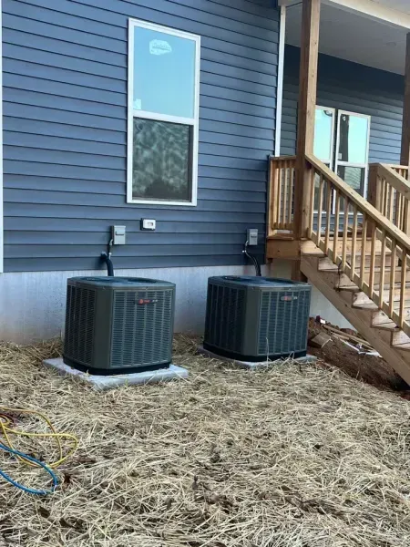 Two gray HVAC units sit on concrete pads against the blue siding of a house next to wooden stairs.
