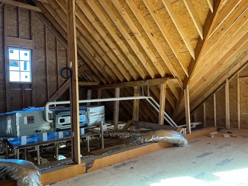 An unfinished attic space with exposed wooden rafters, plywood subflooring, a window, and an HVAC air handler unit.