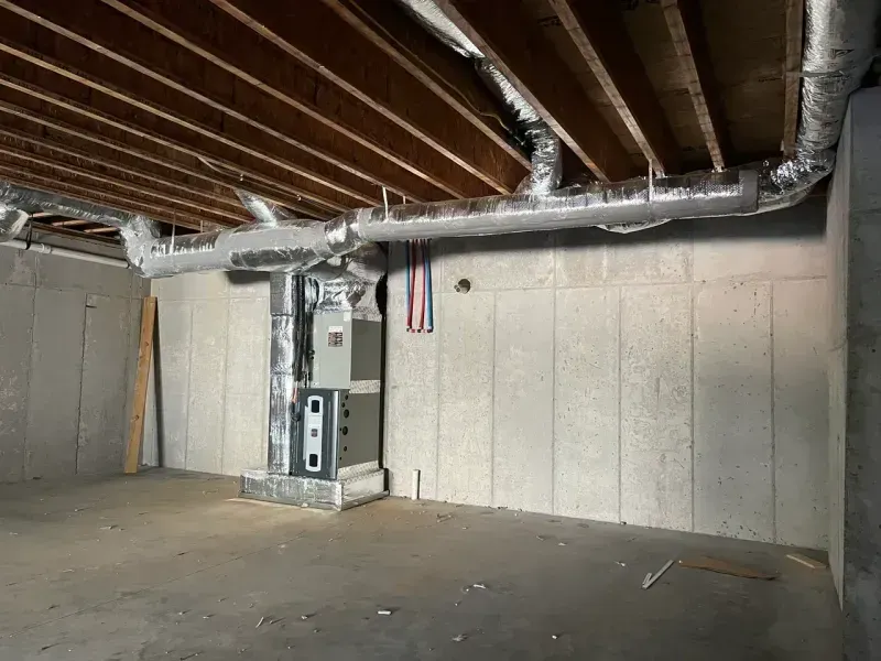 An HVAC furnace unit stands in a bare, unfinished concrete basement with exposed ceiling joists and metallic ductwork.